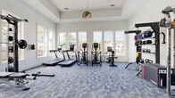 Located at 7326 Wallace Rd in Charlotte, NC, this single-floor fitness center is a comprehensively equipped workout facility designed to support a full range of exercise routines. The space is bathed in natural light, courtesy of large windows lining multiple walls that offer views of the surrounding greenery and neighborhood, creating an open and energizing atmosphere. The interior features soaring ceilings that further enhance the sense of spaciousness, complemented by clean white walls and stylish blue patterned carpet flooring throughout.

The cardio zone is outfitted with treadmills, elliptical machines, and a stationary bike, all positioned to take advantage of the generous window views. A dedicated free weights area includes a full rack of dumbbells in graduated sizes, stability balls, a weight bench, and a power rack with a barbell, making it suitable for both beginner and advanced strength training. A multi-functional cable trainer station provides versatile resistance training options, while additional accessories such as kettlebells, medicine balls, and resistance tools are neatly organized on wall-mounted racks. A built-in shelving unit with a wall-mounted TV and under-counter storage rounds out the space, providing a functional and welcoming hub for members to recharge between sessions.