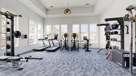 Located at 7326 Wallace Rd in Charlotte, NC, this single-floor fitness center is a comprehensively equipped workout facility designed to support a full range of exercise routines. The space is bathed in natural light, courtesy of large windows lining multiple walls that offer views of the surrounding greenery and neighborhood, creating an open and energizing atmosphere. The interior features soaring ceilings that further enhance the sense of spaciousness, complemented by clean white walls and stylish blue patterned carpet flooring throughout.

The cardio zone is outfitted with treadmills, elliptical machines, and a stationary bike, all positioned to take advantage of the generous window views. A dedicated free weights area includes a full rack of dumbbells in graduated sizes, stability balls, a weight bench, and a power rack with a barbell, making it suitable for both beginner and advanced strength training. A multi-functional cable trainer station provides versatile resistance training options, while additional accessories such as kettlebells, medicine balls, and resistance tools are neatly organized on wall-mounted racks. A built-in shelving unit with a wall-mounted TV and under-counter storage rounds out the space, providing a functional and welcoming hub for members to recharge between sessions.