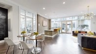 Located at 5350 Burnet Rd in Austin, Texas, this stylish single-floor space seamlessly blends a living lounge and kitchen area into one open, well-appointed layout. The design is unmistakably contemporary, anchored by striking herringbone-patterned flooring that runs throughout and crisp white ceilings equipped with recessed lighting and elegant pendant fixtures.

Floor-to-ceiling windows and full-length glass walls flood the interior with abundant natural light while offering views of an adjacent outdoor patio area, creating a bright and airy atmosphere. Large glass-panel doors provide direct access to the exterior, reinforcing the strong indoor-outdoor connection.

The kitchen counter area features a sleek white bar surface lined with vibrant yellow and teal upholstered bar stools, adding a bold pop of color against the neutral taupe and white walls. A cozy lounge section is anchored by a statement sputnik chandelier overhead, complemented by plush velvet sofas in deep charcoal and grey tones accented with mustard and orange throw pillows. Abstract wall art and geometric wall details add a curated, artistic touch throughout.

A co-working nook with desktop computers is also integrated into the space, alongside glass walls overlooking a well-equipped fitness area. The overall ambiance is polished, welcoming, and energetic — ideal for both relaxing and gathering.