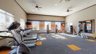 The fitness center at Hurstbourne Estate in Louisville, Kentucky is a generously proportioned, single-floor facility designed to support a full range of exercise routines. The space features soaring ceilings accented with crown molding, dual ceiling fans for air circulation, and recessed lighting throughout, creating a bright and comfortable workout environment. Large windows along multiple walls allow abundant natural light to fill the room while offering views of the surrounding property.

The interior is finished with a durable, patterned carpet in gray and orange tones that complements the room's energetic color scheme — walls are painted in neutral beige with bold orange and blue accent bands running horizontally, reinforcing the athletic atmosphere. The layout is organized to accommodate various workout zones. The cardio area houses a row of treadmills and elliptical machines positioned near the windows. The strength training section includes cable and weight machines, adjustable benches in orange, and a heavy bag stand. A dedicated free weights area offers a full dumbbell rack, stability balls, and wall-mounted mirrors that expand the sense of space. Wall-mounted televisions are positioned for easy viewing during workouts. The facility also includes direct exterior access via a wood-framed glass door, connecting to the broader Hurstbourne Estate community.