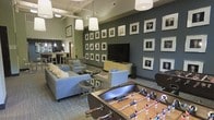 This single-floor amenity lounge is designed to offer both comfort and entertainment in a polished, contemporary setting. The space is anchored by a striking dark accent wall adorned with an extensive gallery of framed black-and-white photographs, creating a sophisticated focal point that draws the eye upward toward the soaring ceilings. Pendant lighting with crisp white drum shades hangs overhead, complementing the industrial-style exposed ceiling elements and contributing to a warm, well-lit atmosphere.

The main seating area features plush blue-gray sofas arranged around sleek glass and metal coffee tables, providing a relaxed environment ideal for socializing. A large flat-screen television mounted on the gallery wall anchors the entertainment zone, making the space equally suited for casual viewing or group gatherings.

Adding a playful dimension to the lounge, a full-size foosball table takes center stage in the foreground, inviting friendly competition among guests. In the background, a bar or kitchenette area with pendant lighting and high seating offers an additional gathering spot, seamlessly integrated into the open floor plan. The overall ambiance blends modern design with inviting warmth, making this a versatile communal space well-suited for relaxation, entertainment, and social interaction.