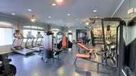This fitness center offers a comprehensive workout environment spread across a single floor. The space is outfitted with a variety of cardio equipment, including treadmills and ellipticals, positioned along the perimeter to take advantage of natural light streaming through arched windows. The strength-training area is well-stocked with multiple cable machines, a multi-function weight station, and adjustable benches with orange upholstery, providing options for a full-body workout. The durable gray flooring is suited for high-traffic use and easy maintenance. Ceiling fans complement the recessed lighting throughout, ensuring the space remains well-ventilated and well-lit. Large mirrors line portions of the walls, enhancing the sense of spaciousness and allowing users to monitor their form. A beverage cooler is visible toward the rear, adding a convenient amenity for hydration. The neutral gray walls and clean layout create a focused, functional atmosphere well-suited for fitness enthusiasts of all levels.