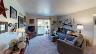 This single-floor residence offers a thoughtfully arranged layout with multiple bedrooms, a living room, dining area, kitchen, and at least one full bathroom. The living room serves as a central gathering space, featuring carpet flooring, a sliding glass door that opens to an outdoor balcony, and a wall-mounted air conditioning unit. Decorative shelving and eclectic wall art contribute to the room's warm, personalized atmosphere.

The dining area accommodates a full-sized wooden table with seating for several, lit by a pendant light overhead. Colorful decorative plates and a mirror accent the walls, adding a homey touch. The kitchen features tile flooring, white cabinetry, standard appliances, and a bold black-and-white buffalo check wainscoting detail that gives the space a distinctive rustic flair.

The primary bedroom is finished with deep olive-green accent walls, carpet flooring, a ceiling fan, and baseboard heating. Natural light enters through multiple windows dressed in white sheer curtains. Additional bedrooms offer varied accent colors, including blue and neutral tones, each with carpeted floors, closets, and ceiling fixtures. The shared bathroom includes a wide vanity with a large mirror and warm wood trim detailing throughout.