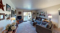 This single-floor residence offers a thoughtfully arranged layout with multiple bedrooms, a living room, dining area, kitchen, and at least one full bathroom. The living room serves as a central gathering space, featuring carpet flooring, a sliding glass door that opens to an outdoor balcony, and a wall-mounted air conditioning unit. Decorative shelving and eclectic wall art contribute to the room's warm, personalized atmosphere.

The dining area accommodates a full-sized wooden table with seating for several, lit by a pendant light overhead. Colorful decorative plates and a mirror accent the walls, adding a homey touch. The kitchen features tile flooring, white cabinetry, standard appliances, and a bold black-and-white buffalo check wainscoting detail that gives the space a distinctive rustic flair.

The primary bedroom is finished with deep olive-green accent walls, carpet flooring, a ceiling fan, and baseboard heating. Natural light enters through multiple windows dressed in white sheer curtains. Additional bedrooms offer varied accent colors, including blue and neutral tones, each with carpeted floors, closets, and ceiling fixtures. The shared bathroom includes a wide vanity with a large mirror and warm wood trim detailing throughout.