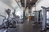 Located at 1440 Ben Sawyer Blvd in Mount Pleasant, South Carolina, this fitness facility at Oyster Park is a single-story space designed to support a comprehensive workout experience. The interior is characterized by soaring vaulted ceilings with exposed ductwork finished in dark tones, contributing to an industrial-modern aesthetic that feels both spacious and functional. Crisp white walls contrast with dark rubber flooring throughout, creating a clean, high-contrast environment well-suited for an active setting.

The cardio zone features a row of Matrix treadmills paired with wall-mounted flat-screen televisions, with a window providing natural light. Nearby, upright stationary bikes and additional cardio equipment offer variety. A dedicated strength training area houses a multi-station cable machine along with free weights displayed on a warm wood dumbbell rack, adding a natural accent to the otherwise sleek palette. Floor-to-ceiling mirrors line portions of the walls, visually expanding the space and supporting proper form during training.

A transitional area near the entrance includes decorative touches such as potted greenery and artwork, softening the utilitarian character of the space. The overall ambiance is modern, clean, and purposefully designed for fitness use.