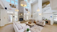 Located at 5489 Crescent Ridge Dr in Dublin, Ohio, this two-story clubhouse at Britton Woods offers a refined and welcoming environment across two well-appointed floors. The main level greets visitors with a bright entryway featuring hardwood-style flooring, glass-paneled French doors, and an abundance of natural light. The heart of this floor is a dramatic double-height great room anchored by a soaring stone fireplace that stretches toward the ceiling, flanked by large windows that fill the space with daylight. Elegant pendant lighting, white columns, and a classic staircase with white balusters enhance the architectural character of the room. A curved, bar-height kitchen island with granite countertops serves as the centerpiece of the well-equipped kitchen, complemented by white cabinetry, hardwood-style flooring, and generous counter space — ideal for hosting and gatherings. A casual lounge area with a wall-mounted TV and high-top dining table provides a relaxed setting for smaller groups. The upper level features an open-air hallway with white railings overlooking the grand living area below, illuminated by hanging lantern-style fixtures and exposed beam detailing. Throughout, the space maintains a cohesive palette of whites, neutrals, and warm gold accents, creating an atmosphere that is both sophisticated and inviting.