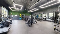 Located at 600 Bond St in Bridgeport, Connecticut, this contemporary fitness center offers a generously sized, single-floor exercise space designed to accommodate a wide range of workout needs. The open-plan layout is defined by an exposed industrial ceiling fitted with striking rectangular LED light fixtures that cast bright, even illumination throughout the room, creating an energizing atmosphere.

The cardio zone is anchored by large floor-to-ceiling windows that frame lush outdoor greenery, flooding the space with natural light and providing a refreshing view during workouts. Equipment in this area includes treadmills, ellipticals, a stair climber, spin bikes, and rowing machines, all arranged on durable rubber flooring.

The strength training area features a comprehensive selection of cable machines, multi-functional weight stations, air bikes, and adjustable benches, complemented by full-length mirrors that enhance the sense of space. A dedicated free weights section provides a complete dumbbell rack alongside additional benches.

A distinct functional training zone is set apart by dramatic faux-greenery accent walls that climb to the ceiling, adding a vibrant, organic element to the otherwise industrial aesthetic. This area is equipped with a heavy punching bag, battle ropes, a squat rack, and resistance tools. Together, these zones create a well-rounded, modern facility suited for diverse fitness goals.