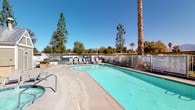This single-story residential community offers a thoughtfully designed set of shared amenities that cater to everyday convenience and outdoor enjoyment. At the heart of the outdoor space is a generously sized swimming pool with clear turquoise water, surrounded by a concrete deck lined with lounge chairs — ideal for relaxing under the sunny skies. Tall palm trees and mature pines frame the pool area, while distant mountain views provide a picturesque natural backdrop visible beyond the white perimeter fencing. The grounds are well-maintained, with open walkways connecting the various areas of the property. A covered mailbox station serves as a central hub for residents, housing a large bank of individual, labeled U.S. Mail compartments neatly mounted along the wall, alongside an outgoing mail slot for added convenience. The covered alcove provides shade and shelter, keeping the area protected from the elements. The community's exterior features a neutral tan siding palette consistent throughout the buildings, contributing to a cohesive and clean aesthetic. Landscaping with shrubs and greenery adds a touch of nature to the pathways. Overall, the community presents a functional and comfortable environment with key amenities easily accessible to all residents.