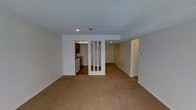 This single-floor space offers a practical, open layout designed for comfortable everyday living. The main living area is finished with wall-to-wall beige carpeting, creating a warm and cohesive feel throughout. Neutral-toned walls and a textured ceiling contribute to a clean, understated aesthetic.

A defining architectural detail is the partial white divider with vertical slat openings, which subtly separates the living area from the adjoining spaces while maintaining an open, connected feel. This divider frames the view into the kitchen, which features wood-toned cabinetry, a gas range with overhead ventilation, and hard flooring — a functional contrast to the carpeted living zones.

Recessed lighting is installed in key areas, providing consistent illumination across the space. A central hallway extends toward the back of the unit, leading to additional rooms, each accessible through white panel doors. A second doorway on the right side of the main living area suggests further rooms or utility spaces beyond.

Overall, the layout is straightforward and efficiently organized, making it well-suited for a variety of uses. The neutral finishes throughout provide a versatile backdrop adaptable to different furnishing styles.