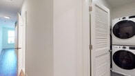 This well-appointed single-floor space offers a clean, modern aesthetic defined by warm hardwood-style flooring and crisp white walls that run throughout. The layout, as revealed in the floorplan view, is thoughtfully organized with distinct living, sleeping, and utility areas flowing in a linear arrangement.

The kitchen is a standout feature, equipped with stainless steel appliances including a refrigerator, range, over-the-range microwave, and dishwasher. White shaker-style cabinetry reaches toward the ceiling for ample storage, while a textured penny-tile backsplash adds a stylish focal point. Light-toned countertops and a single-basin sink complete the functional yet attractive cooking space.

The living and sleeping areas are generously proportioned, bathed in natural light from multiple windows fitted with horizontal blinds. The hardwood-style flooring continues seamlessly through these rooms, enhancing the sense of openness and continuity.

Storage is well considered, with a walk-in closet outfitted with wire shelving systems offering organized space for clothing and essentials. Louvered bifold doors are a consistent detail throughout, adding a classic touch to the overall design.

A dedicated laundry area with a washer and dryer rounds out the practical amenities of this space. The overall ambiance is bright, clean, and move-in ready, with modern finishes that create a welcoming and functional environment.