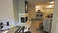 This single-floor space showcases a thoughtfully designed open-concept layout that seamlessly connects the living and kitchen areas. The living space is anchored by a striking corner fireplace with a crisp white surround, topped by an ornate black-framed mirror that adds a decorative focal point to the room. Wood-look flooring runs throughout, lending warmth and visual continuity across the space.

The neutral gray wall tones create a versatile backdrop, complemented by a cowhide-style area rug that adds texture and contrast to the living area. A glass-top coffee table and a decorative barrel-style accent stool contribute to the eclectic yet cohesive design aesthetic.

The kitchen, visible from the living area, features white cabinetry, a dark mosaic tile backsplash, and a breakfast bar with wrought iron barstools, providing both a casual dining option and a natural boundary between the two spaces. Overhead track lighting in the kitchen ensures the area is well-lit. The open sightlines between rooms enhance the sense of space, making the overall layout feel connected and functional.