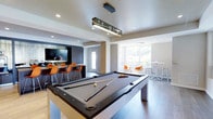 Located at 1 Cotton St in Philadelphia's Manayunk neighborhood, this single-floor amenity space is designed to serve as a dynamic social hub for its community. The open-plan layout is anchored by a sleek, contemporary billiards table positioned beneath a striking rectangular pendant light fixture, creating an immediate focal point upon entry. The flooring throughout combines light-toned tile and wood-style planks, lending a clean, modern aesthetic to the entire space. Adjacent to the billiards area, a dedicated media lounge features a large wall-mounted flat-screen television framed by dark cabinetry, paired with a row of vibrant orange bar-style chairs that add a bold pop of color against the neutral palette. Recessed lighting complements the pendant fixtures overhead, ensuring the space is well-lit throughout. On the opposite end, a bright and airy seating nook benefits from generous natural light streaming through large windows, furnished with contemporary white and orange accent chairs alongside high-top tables — ideal for casual conversation or individual use. The overall ambiance strikes a balance between energetic and inviting, making it a versatile venue suited for both lively social events and relaxed everyday use.