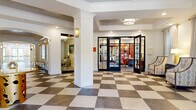 Located at 137 Roseberry Rd in Columbia, SC, this sophisticated two-story clubhouse offers a generously proportioned community gathering space designed with upscale finishes throughout. The main floor welcomes guests with a striking checkered stone-tile foyer framed by white columns, tray ceilings, and recessed lighting. A glass-paned French door entry leads into a bright business center accented with bold coral and red tones, while an adjacent lounge area features cream sofas, vibrant floral rugs, gold cage pendant lighting, and a built-in mail center. A beverage bar with stone countertops and a wine cooler adds a convenient amenity nearby. The upper floor dramatically expands the space, revealing a soaring double-height great room with rich herringbone hardwood floors, floor-to-ceiling wainscoting panels, a marble fireplace, and sunburst gold flush-mount fixtures overhead. A sleek glass-and-steel staircase railing connects the two levels with modern elegance. A formal dining room on this level showcases deep navy board-and-batten accent walls, a rustic wood dining table, oval upholstered chairs, and a statement geometric brass chandelier. Large windows throughout invite natural light, while thoughtfully curated décor creates a refined yet welcoming ambiance.