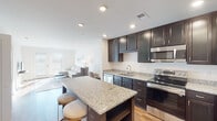 This single-floor residence offers a thoughtfully designed open-concept layout that seamlessly connects the kitchen, dining, and living areas. The kitchen is a standout feature, showcasing rich dark cabinetry, granite countertops, a center island with bar seating, and a full suite of stainless steel appliances including a refrigerator, electric range, microwave, and dishwasher. Warm hardwood-style flooring flows throughout the main living areas, complementing the bright, neutral color palette and recessed lighting overhead. The living room is bathed in natural light from large windows and a glass door that opens to the exterior, creating an airy and inviting atmosphere. The bedrooms are comfortably sized, carpeted for warmth, and feature windows that bring in ample daylight. The bathroom is clean and modern, appointed with dark cabinetry, a white countertop vanity, a large mirror, and classic black-and-white tile flooring with a shower/tub combination. A dedicated laundry area with a stacked washer and dryer, along with built-in wire shelving for additional storage, adds significant practicality to the unit. Overall, the space strikes a balance between modern finishes and functional living.