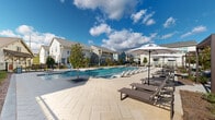 Located at 1001 NW Cernan Dr in Huntsville, Alabama, this outdoor community amenity area showcases a thoughtfully designed resort-style pool complex. The centerpiece is a generously sized swimming pool with crystal-clear blue water, featuring an accessible entry lift, stainless steel entry rails, and a shallow wading ledge that extends into the pool. The surrounding deck is finished with large-format stone pavers, providing ample space for relaxation. Rows of cushioned chaise lounges and dark-framed lounge chairs are arranged along the poolside, complemented by large shade umbrellas for comfort under the Alabama sun. Raised planter beds featuring ornamental trees with fire-pit style bases add a refined landscaping detail throughout the deck area. The pool area is enclosed by a sleek black metal fence, ensuring safety while maintaining open sightlines. Surrounding the pool are several charming buildings rendered in a modern farmhouse architectural style, featuring board-and-batten siding, metal roofing, large multi-pane windows, and red accent doors. These structures appear to serve as clubhouse and amenity facilities for the community. The overall ambiance is bright, welcoming, and well-appointed, offering residents a polished outdoor retreat.