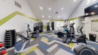 Located at 101 Arbor Run Dr in Lincolnton, NC, this fitness center offers a well-appointed exercise space designed to support a comprehensive workout routine. The room is defined by striking vaulted ceilings that create an open, airy atmosphere, complemented by recessed lighting and multiple windows that invite abundant natural light. A bold green accent stripe runs along the walls, adding a lively, energetic aesthetic that is further enhanced by the eye-catching chevron-patterned carpet in shades of blue, green, yellow, and gray.

The space is equipped with a diverse selection of cardio machines, including treadmills, elliptical trainers, and a stationary bike, providing ample options for aerobic exercise. Strength training is well represented with a multi-station cable machine, a weight bench, and additional free weight accessories. Built-in shelving along one wall offers convenient storage, while a wall-mounted flat-screen TV keeps users entertained during workouts.

A vending machine is available for refreshments, and a small seating area with a table and chairs provides a spot to rest or review workout plans. French doors on one end of the room open to an outdoor area, further enhancing the sense of space and connectivity to the outdoors. The overall design is modern, functional, and energizing.
