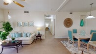 This single-floor residence is designed with a cohesive coastal aesthetic, blending soft aqua and neutral tones throughout. The open living area serves as the heart of the home, featuring light wood-style flooring, a ceiling fan, and a striking arched fireplace with a tiled surround. A large sliding glass door floods the space with natural light and provides direct access to a private patio overlooking a landscaped outdoor area. Decorative touches — including a woven sunburst wall accent and framed coastal artwork — reinforce the seaside ambiance.

The dining area sits adjacent to the living space, accommodating a full dining set with aqua upholstered chairs. The kitchen is well-appointed with light wood cabinetry, granite-style countertops, stainless steel appliances including a range, microwave, refrigerator, and dishwasher, plus an arched window above the sink that brings in additional natural light.

The primary bedroom offers a serene retreat with plush carpeting and framed shell artwork above the bed. A secondary room features a charming arched window and daybed, ideal as a guest space or sitting room. The bathroom includes a full tub and shower combination with clean white tile. A hallway connects the private quarters, completing this well-organized and inviting floor plan.