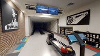 The Parkway Lofts bowling lounge is a thoughtfully designed recreational amenity that blends retro aesthetics with modern functionality. The space centers around two full-length bowling lanes equipped with automatic ball returns and digital touchscreen scoring systems, with overhead monitors displaying lane assignments and welcoming residents by name. A dedicated ball rack along the lane perimeter keeps a colorful selection of bowling balls readily accessible.

The flooring throughout the lane area features a striking patchwork of multicolored carpet tiles in red, blue, yellow, and gray, reinforcing the playful, retro atmosphere. Exposed brick walls line portions of the space, lending an industrial warmth that contrasts with the clean white walls elsewhere. Bold vintage-style bowling graphics and wall art, including retro promotional prints, are mounted throughout, adding personality and visual interest.

Adjacent to the lanes, a generously sized lounge area provides comfortable seating for spectators and groups, with a large wraparound turquoise sectional sofa set against the exposed brick backdrop and coffee tables positioned for casual socializing. A shoe rental cubby and entry door with natural light complete the functional layout. Recessed lighting and large circular ceiling fixtures maintain a well-lit, welcoming ambiance throughout the entire space.