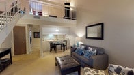 This two-story residence in the 63021 area of Missouri offers a thoughtfully designed layout across two levels. The main living area immediately impresses with soaring vaulted ceilings that extend upward to an open loft above, accessible via a staircase and framed by a metal railing. The living room provides a generous, open feel with plush carpeting and seamless flow into the dining area, which is illuminated by a pendant light and positioned adjacent to the kitchen. The galley-style kitchen features warm wood cabinetry lining both walls, white countertops, a full suite of appliances including a dishwasher and range, and tile flooring. The upper loft level serves as a comfortable lounge or sitting area with angled ceilings, soft carpet, and ample room for relaxed seating. Two well-proportioned bedrooms are each carpeted and filled with natural light from large windows with blinds. One bedroom includes a spacious walk-in closet with built-in shelving. Each bathroom is distinctly appointed — one with a shower/tub combination and warm wood vanity cabinetry, and the other featuring a standalone glass-enclosed shower stall. Throughout, neutral tones and warm lighting create a cohesive and inviting ambiance.