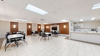 Located at 2470 Southvale Crescent in Ottawa, this single-floor multi-purpose space offers a generously sized, open-concept layout well-suited for gatherings, meetings, or community events. The room is finished with large-format white ceramic floor tiles featuring a subtle geometric pattern, lending a clean and contemporary aesthetic throughout.

The main area is furnished with a combination of round and rectangular white tables paired with black chairs, accommodating a variety of seating arrangements. A warm accent wall clad in rich wood paneling anchors one side of the room and frames a wall-mounted flat-screen display, making the space functional for presentations or events. Wall sconces provide soft, ambient lighting alongside a suspended drop-tile ceiling fitted with flush skylight-style light panels that brighten the interior evenly.

A kitchenette area along one wall includes white cabinetry, a countertop with sink, refrigerator, and microwave, supporting catering or refreshment needs. The space is also equipped with a wall-mounted split air conditioning unit for climate control. A whiteboard is available for collaborative use. Access is provided through a glass-panel door, and the layout allows for flexible furniture configurations to suit diverse functions.
