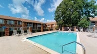 This single-story property showcases a well-maintained outdoor amenity area centered around a spacious swimming pool with clear blue water. The pool deck is finished in light-toned concrete and is furnished with lounge chairs, providing a comfortable setting for outdoor relaxation. Entry to the pool is facilitated by built-in metal handrails on at least one side, and safety equipment is visibly mounted nearby.

The surrounding building is constructed of classic red brick with a warm brown roof, lending the property a traditional and durable aesthetic. Covered balconies with white railings line the building's exterior, offering shaded outdoor spaces that overlook the pool area. A wrought iron-style perimeter fence encloses the pool area, providing both security and a clean visual boundary.

Mature, leafy trees frame the background, adding natural shade and greenery to the outdoor environment. The overall ambiance is relaxed and residential, with an open-air feel enhanced by the surrounding landscaping. The space benefits from ample natural light, making the pool area an inviting outdoor retreat under open skies.