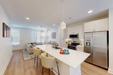 This well-designed three-story home offers a thoughtful layout across three distinct levels, delivering efficient and functional living spaces throughout. The ground floor houses a garage, utility room, and entrance, providing a practical base for the home. The main living level is anchored by a bright, open-concept space that seamlessly combines the kitchen, dining, and living areas. The kitchen features white shaker-style cabinetry, stainless steel appliances including a French door refrigerator, a large waterfall-edge island with bar seating, and pendant lighting that adds a modern touch. The adjacent dining area benefits from natural light streaming through French doors, while the living room offers a relaxed, airy atmosphere with large windows. The upper floor hosts two bedrooms, including a primary suite with an en-suite bathroom featuring a white floating vanity with a black-framed mirror and marble-style tile flooring. Walk-in closets with built-in shelving provide generous storage. The secondary bedroom is well-proportioned with a built-in closet and ceiling fan. A dedicated office and laundry room add further practicality. Throughout, light hardwood flooring, a neutral color palette, and recessed lighting create a clean, contemporary ambiance.