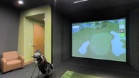 This single-floor indoor golf simulator space is purpose-built for an immersive and focused golf experience. The room is finished with dark, textured acoustic wall panels on all sides, creating an environment that minimizes distractions and reduces sound reflection — ideal for both practice and simulated play. At the far end of the room, a large, wide-format projection screen displays high-resolution golf course visuals, bringing realistic course environments to life. The simulation system showcases detailed aerial and ground-level views of fairways, greens, and bunkers, providing an authentic and engaging gameplay experience. A section of artificial turf serves as the hitting mat, positioned directly in front of the screen to replicate the feel of a real fairway or tee box. A golf bag with a full set of clubs stands ready near the entrance, underscoring the room's functional design. The entry door features a warm wood-toned finish, offering a subtle contrast to the room's otherwise dark interior. The space is efficiently laid out to maximize the swing area while keeping all necessary equipment within easy reach, making it a well-equipped and practical facility for golfers of all skill levels.