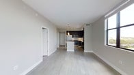 This single-floor space showcases a contemporary open-concept design that seamlessly connects the living area and kitchen. Light gray hardwood flooring flows throughout, contributing to a clean, airy aesthetic complemented by neutral-toned walls and crisp white baseboards.

The kitchen is a standout feature, equipped with dark espresso cabinetry that creates a striking contrast against the light surroundings. Stainless steel appliances, including a refrigerator, microwave, and range, provide a polished, modern finish. A white kitchen island anchors the space, offering additional prep and seating potential. Pendant lighting hangs above the island, adding a refined touch, while recessed track lighting ensures the kitchen is well-illuminated.

The open living area is generously proportioned, offering flexible furniture arrangement options. Visible HVAC vents integrated into the ceiling maintain a streamlined look. A doorway off the main living space leads to additional rooms, and a white entry door is visible in the background, reinforcing the unit's bright, welcoming atmosphere. Overall, the space presents a well-designed, move-in-ready interior with a cohesive modern aesthetic.