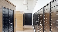 This dedicated mailroom offers a thoughtfully designed space for mail and package management within a residential community. The room is organized around a series of freestanding wood-clad kiosks and wall-mounted units, all finished in light natural oak-toned paneling that lends warmth and a contemporary aesthetic to the space.

The perimeter and central kiosks house an extensive array of individual mailboxes with brushed metal hardware, providing ample mailbox capacity for a large number of residents. Alongside the traditional mailboxes, a full bank of stainless steel package lockers lines one corridor, offering secure, automated parcel storage for convenient 24/7 package retrieval.

The room benefits from elevated ceilings with clerestory windows that admit generous natural light, creating an airy and open atmosphere. A series of pendant lantern-style light fixtures hang at varying heights, providing both functional illumination and a refined design touch. The light-toned hardwood-style flooring complements the wood paneling throughout, maintaining a cohesive and polished interior palette.

A subtle geometric-patterned accent wall adds visual interest, while the overall layout ensures clear, efficient circulation between the various mailbox and locker stations. Stainless steel waste receptacles are conveniently positioned throughout the space. The mailroom's design balances practicality with elevated finishes, reflecting a high standard of communal amenity design.