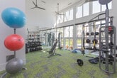 Located at 2789 Satellite Blvd in Duluth, GA, this well-appointed fitness center is designed to support a full range of workout styles and training needs. The space is defined by its soaring double-height ceilings and expansive floor-to-ceiling windows along one wall, which flood the interior with natural light and offer views of the surrounding landscaped grounds. Ceiling fans circulate air throughout, contributing to a comfortable workout environment.

The fitness center is equipped with an extensive selection of free weights, including a full dumbbell rack, weight plates, and barbells. A multi-functional cable and weight machine supports a variety of strength training exercises, while a dedicated functional training rig features TRX suspension straps, resistance bands, medicine balls, and kettlebells for dynamic, full-body workouts. An adjustable weight bench and a leg extension/curl machine round out the strength training offerings.

Full-length mirrors line one wall, enhancing the sense of space while allowing users to monitor their form. The vivid green and grey patterned carpet tile flooring adds a modern, energetic aesthetic to the space. An adjacent room, accessible through frosted glass sliding doors, provides additional space and includes a wall-mounted television, offering a more private area for stretching or alternative training activities.