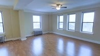 This single-floor space presents a clean, open layout highlighted by warm hardwood flooring that reflects abundant natural light. The main living area is defined by three large windows with classic black frames, providing expansive views of surrounding trees and filling the room with daylight. Crown molding runs along the ceiling's perimeter, adding a traditional architectural detail that lends the space a polished character. A ceiling fan with a centered light fixture serves as a functional focal point overhead. A cast-iron radiator along one wall is indicative of classic building construction, offering a charming, vintage detail while providing efficient heating. The walls are painted in a neutral, warm beige tone, creating a versatile backdrop suited to a variety of interior styles. White baseboards offer a crisp contrast against both the walls and flooring. The generous proportions of the room allow for flexible furniture arrangements, making it well-suited for comfortable living. Overall, the space combines classic architectural elements with a bright, airy ambiance that feels both welcoming and functional.