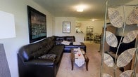 This single-floor residence offers an open-concept layout that seamlessly connects the main living areas, creating a cohesive and functional living environment. The living room serves as the heart of the space, featuring plush carpeted flooring and a large, dark leather sectional sofa that provides ample seating. A wooden coffee table anchors the seating area, while a large framed cityscape artwork adds a bold decorative focal point on the wall. The room is illuminated by a ceiling-mounted light fixture, and a textured ceiling adds subtle character overhead. The open floor plan allows direct sightlines into the dining area, which accommodates a dining table and chairs, ideal for everyday meals. Beyond the dining space, the kitchen is visible with light-colored cabinetry, a countertop bar area, and standard appliances including a refrigerator and microwave. A shelving unit near the dining area provides additional storage and display space. The overall ambiance of the space is warm and inviting, with a practical layout that maximizes the flow between living, dining, and cooking areas, making it well-suited for comfortable day-to-day living.