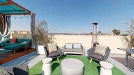 This rooftop terrace is a thoughtfully designed outdoor retreat that blends comfort, style, and functionality. The space is anchored by two elegantly appointed cabana structures, featuring rich wood-framed pergolas draped with flowing striped curtains and vibrant teal canopies. Pendant string lights add a warm ambiance, while the raised hardwood deck platform beneath the cabanas hosts both a dining area and a plush lounge seating arrangement with colorful accent pillows.

Adjacent to the cabanas, an artificial turf lounge zone provides a relaxed gathering spot, furnished with woven outdoor chairs, a loveseat, cylindrical concrete coffee tables, and patio side tables. A tall pyramid patio heater ensures year-round usability, and a potted olive tree adds a touch of natural greenery.

A covered colonnade runs along one side of the rooftop, framed by classical white columns, and houses a ping-pong table for recreational use. This corridor also provides access to a built-in outdoor kitchen and connects to the main corridor access point, which is equipped with a fire extinguisher station.

Throughout the terrace, sweeping panoramic views of the surrounding cityscape, distant mountains, and palm trees are visible under open blue skies, making this rooftop an exceptional venue for both social gatherings and quiet relaxation.
