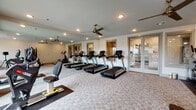 This fitness center is a thoughtfully designed, single-floor facility offering a comprehensive workout environment. The space is outfitted with a row of treadmills, elliptical trainers, and a seated rowing machine, providing a full range of cardio options. A dedicated strength training area includes a multi-function cable machine, an adjustable weight bench, and a dumbbell rack with a complete set of free weights. The flooring throughout consists of durable, patterned carpet tiles well-suited for high-traffic fitness use, with rubber mats placed beneath equipment for added stability and protection. A full-length mirror spans one wall, enhancing the sense of openness and allowing users to monitor their form during workouts. Recessed lighting provides consistent illumination across the space, complemented by multiple ceiling fans that promote air circulation. Large windows along one wall invite abundant natural light and offer views to the exterior. Glass-paneled doors connect to an adjacent area, adding to the open, airy feel of the facility. The overall ambiance is clean, modern, and functional, offering a well-rounded fitness experience in a comfortable and well-maintained environment.