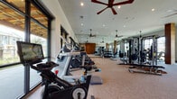 This well-appointed fitness center offers a thorough and thoughtfully designed workout environment spread across a single open floor. The space is characterized by soaring ceilings fitted with multiple large ceiling fans, recessed lighting, and expansive floor-to-ceiling windows and glass walls that flood the interior with abundant natural light while framing views of the surrounding outdoor landscape.

The cardio zone is generously stocked with a lineup of FreeMotion cycling bikes, Precor ellipticals, and multiple treadmills, all arranged to take advantage of the natural light and outdoor views. Adjacent to this area, a dedicated strength training section features Axis cable machines, a power rack, weight benches, kettlebells, and battle ropes, providing a full complement of resistance training options.

Full-length mirrors line portions of the walls, enhancing the sense of spaciousness and supporting proper form during workouts. Warm wood accent columns punctuate the interior, adding a natural, contemporary aesthetic that balances the modern equipment. The neutral-toned carpeted flooring ties the space together while providing comfort underfoot. The overall ambiance is bright, open, and motivating — a premium facility designed to support a wide range of fitness goals.