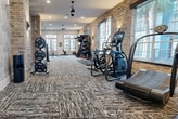 This state-of-the-art fitness facility, located at 3210 Louisiana in Houston, TX, spans an impressive 2,340 square feet on a single floor. The space is thoughtfully designed to accommodate various workout styles and preferences. 

Upon entry, visitors are greeted by a stylish lobby area featuring a unique vertical garden wall, adding a refreshing touch of nature to the urban setting. The flooring throughout is a distinctive patterned carpet, providing both aesthetic appeal and functionality.

The main workout area is divided into several sections, each dedicated to specific exercise types. A spacious cardio zone houses multiple treadmills, ellipticals, and stationary bikes, all facing large windows that allow natural light to flood the space. The strength training area features an array of weight machines and free weights, complemented by exposed brick walls that lend an industrial chic vibe.

A notable architectural feature is the central brick column that serves both structural and aesthetic purposes, effectively dividing the space while maintaining an open feel. Ceiling-mounted fans and recessed lighting fixtures ensure comfort and proper illumination throughout.

The facility also includes a specialized functional training area with equipment like medicine balls, resistance bands, and a multi-functional fitness rig. A striking circular staircase with ornate iron railings adds a touch of elegance, possibly leading to additional spaces or offices.

Overall, this fitness center combines modern amenities with thoughtful design elements, creating an inviting and motivating environment for health enthusiasts.