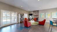 Located at Willow Creek in Tempe, Arizona, this community living room is a thoughtfully designed single-floor space that balances comfort and functionality with a bold, contemporary aesthetic. The room features large-format dark tile flooring throughout, complemented by neutral gray walls that serve as a backdrop for vibrant pops of color in the furnishings and artwork.

A dramatic floor-to-ceiling stacked stone fireplace wall anchors one side of the room, paired with a wall-mounted flat-screen TV and rustic wood shelving — creating an inviting focal point for relaxation. A cluster of bold red leather lounge chairs arranged around a round white coffee table atop a plush green area rug defines the primary seating zone.

On the opposite side, the space opens into a casual dining area with a round table, cylindrical striped ottomans, and pendant lighting above. A white credenza with a decorative sunburst mirror and small appliances serves as a refreshment station, while open wood shelving units display books and décor. French doors and large windows allow natural light to fill the space and offer views to the outdoors. Recessed lighting and pendant fixtures provide warm, layered illumination throughout, completing this welcoming and multifunctional community gathering area.