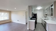 This single-floor space showcases a thoughtfully designed interior that blends contemporary style with functional living. The kitchen is a standout feature, equipped with stainless steel appliances, including a refrigerator and gas range, complemented by sleek gray shaker-style cabinetry that extends to the upper walls for ample storage. A mosaic tile backsplash in soft blue and white tones adds a decorative touch, while the crisp white countertops provide a clean contrast against the darker appliances. Recessed and flush-mount ceiling lighting ensures the kitchen is well-lit throughout.

The space transitions smoothly into an open living area, characterized by neutral white walls adorned with classic crown molding, adding an elegant architectural detail. The dark wood-look flooring runs consistently throughout, providing a warm and cohesive aesthetic. White baseboards and interior doors with raised panel detailing contribute to the clean, polished look of the space. A structural column elegantly divides the kitchen from the living area, maintaining a sense of openness while defining each zone. Overall, the space presents a modern yet timeless ambiance, ideal for comfortable everyday living.