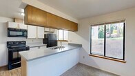 Located in Fircrest, Washington, this single-floor, 2-bedroom, 2-bathroom residence offers a practical and well-organized layout. The living and dining area is generously sized, featuring soft carpet flooring, a textured ceiling, and large sliding glass doors that flood the space with natural light and provide views of the surrounding greenery. Baseboard heating is present throughout, ensuring year-round comfort.

The kitchen is equipped with a peninsula counter featuring a gray laminate surface and an integrated sink, complemented by warm wood-toned upper cabinetry and white lower cabinets. Black appliances, including a refrigerator, range, and microwave, provide a cohesive look, while a window above the counter brings in additional natural light.

The primary bedroom is a comfortable retreat with plush carpeting, a closet, and an en-suite bathroom connection. The second bedroom also features carpet flooring, a baseboard heater, and a bright window with outdoor views. Both bathrooms are functional spaces; one includes patterned wallpaper, wood-look flooring, a vanity with under-sink storage, and a tub-shower combination. The entry area features wood-look flooring and a closet, providing a welcoming transition into the home.