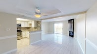 This single-floor space offers a seamlessly connected open-plan layout, combining the kitchen and living area into one bright, airy environment. The kitchen is well-appointed with white cabinetry, stainless steel appliances, and granite countertops, complemented by a striking mosaic tile backsplash that adds a modern design accent. A peninsula-style counter provides a natural division between the kitchen and the living space while maintaining the open feel of the floor plan. The living area is finished with large-format, glossy white tile flooring that reflects natural light throughout the space, enhancing the sense of openness. A ceiling fan with an integrated light fixture is centrally positioned above a subtle tray ceiling detail, adding architectural interest overhead. Large sliding glass doors at the far end of the living area invite abundant natural light and provide views to the outdoors, creating a seamless connection between the interior and exterior. Neutral wall tones throughout the space contribute to a clean, versatile aesthetic that suits a variety of interior styles. Overall, the space presents a functional and modern single-floor layout with thoughtful finishes and a bright, welcoming ambiance.