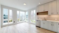 This contemporary single-floor residence showcases a clean, modern design throughout, unified by light-toned hardwood flooring and a crisp white palette. The open-concept living and kitchen area is anchored by a striking corner window configuration that floods the space with natural light and frames impressive city skyline views. The kitchen is thoughtfully equipped with stainless steel appliances, including a refrigerator, range, microwave, and dishwasher, complemented by sleek white cabinetry, warm wood accent shelving, a tile backsplash, and light countertops. The layout connects the kitchen and living areas seamlessly, creating an inviting, functional environment. Two well-proportioned bedrooms offer a calm retreat, featuring the same warm hardwood flooring, soft neutral wall tones, and baseboard heating. The bathroom is finished to a high standard, with a wide single-basin vanity, a backlit mirror, clean white cabinetry, and a glass-enclosed shower. A second vanity area provides additional convenience. Throughout the unit, recessed and flush-mount lighting ensures a bright, welcoming ambiance, while the consistent use of modern finishes contributes to a cohesive and polished aesthetic.