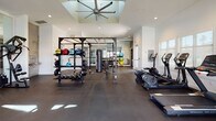 This single-story fitness center is thoughtfully designed to accommodate a wide range of workout styles across several distinct zones. The main strength and functional training area is generously proportioned, featuring dark rubber flooring, a power rack loaded with kettlebells, medicine balls, and a heavy bag, alongside a full dumbbell rack. A large ceiling fan mounted above a recessed skylight keeps the space well-ventilated and bright. Cardio equipment, including rowing machines and ellipticals, lines one side of this area.

A dedicated treadmill alcove is positioned adjacent to a full-height glass garage door, allowing natural light and outdoor views of the surrounding landscaping to flood the space during workouts. The strength machine zone features Promaxima cable and resistance equipment beneath vaulted ceilings and large windows that further enhance the airy atmosphere.

The cycling studio is a standout feature, housing a row of Pro Maxima spin bikes facing a large wall-mounted flat-screen TV, all reflected by a full floor-to-ceiling mirror that amplifies the sense of space. The entry corridor is finished with decorative hexagonal metallic tile surrounding a dual-level water bottle filling station, adding a polished aesthetic touch near the clearly posted fitness center rules and glass entry door.