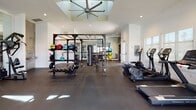 This single-story fitness center is thoughtfully designed to accommodate a wide range of workout styles across several distinct zones. The main strength and functional training area is generously proportioned, featuring dark rubber flooring, a power rack loaded with kettlebells, medicine balls, and a heavy bag, alongside a full dumbbell rack. A large ceiling fan mounted above a recessed skylight keeps the space well-ventilated and bright. Cardio equipment, including rowing machines and ellipticals, lines one side of this area.

A dedicated treadmill alcove is positioned adjacent to a full-height glass garage door, allowing natural light and outdoor views of the surrounding landscaping to flood the space during workouts. The strength machine zone features Promaxima cable and resistance equipment beneath vaulted ceilings and large windows that further enhance the airy atmosphere.

The cycling studio is a standout feature, housing a row of Pro Maxima spin bikes facing a large wall-mounted flat-screen TV, all reflected by a full floor-to-ceiling mirror that amplifies the sense of space. The entry corridor is finished with decorative hexagonal metallic tile surrounding a dual-level water bottle filling station, adding a polished aesthetic touch near the clearly posted fitness center rules and glass entry door.