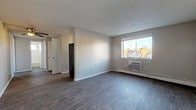 This single-floor unit offers a straightforward, functional layout spread across several distinct rooms. The main living area is finished with wood-style luxury vinyl flooring in a warm gray tone, neutral-painted walls, and a textured ceiling. A wall-mounted air conditioning unit and a baseboard heater provide year-round climate control, while a window offers natural light and exterior views. The open layout connects to additional rooms via a central hallway.

The bedrooms are carpeted for comfort and each feature baseboard heating and windows that bring in natural light. One bedroom includes a sliding-door closet with a rod and shelf system, while another features built-in open shelving and a closet area, providing practical storage solutions. Both rooms maintain the same neutral color palette seen throughout the unit.

The bathroom is finished with wood-look vinyl flooring, white tile surrounding a full soaking tub with a shower, a vanity with dark countertops, and a small window for ventilation. An additional area near the entry features hard flooring and a baseboard heater. Throughout the unit, the consistent use of neutral tones, hard-surface and carpeted flooring, and ample baseboard heating contributes to a clean, practical living environment.