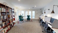 This thoughtfully designed single-floor workspace combines functionality with contemporary style. The open layout is anchored by plush, high-back lounge chairs in rich teal upholstery arranged around a sleek round table, creating an inviting collaborative zone ideal for informal meetings or focused work. Complementing this seating area, a row of dedicated workstations lines the right wall, each equipped with ergonomic task chairs and a continuous built-in desk surface, offering a structured environment for heads-down productivity.

Natural light filters generously through a series of large windows fitted with roller shades along the back wall, brightening the neutral, cream-toned walls and enhancing the airy feel of the space. Recessed ceiling lighting provides consistent, even illumination throughout. The carpeted floor adds warmth and acoustic comfort to the room.

A built-in bookshelf stocked with books anchors one corner, contributing an intellectual, creative atmosphere. A striking geometric wire wall sculpture serves as a focal art piece, adding visual interest and personality. The overall ambiance is clean, modern, and professionally curated to support both collaborative interaction and individual focus.
