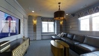 Located at 722 Island Park Drive on Daniel Island, South Carolina, this single-floor space showcases a thoughtfully curated interior defined by a blend of classic architectural elements and contemporary design. The main living area immediately draws attention with its distinctive horizontal shiplap-style wall paneling, painted in a soft grey-blue tone that lends the room a calm, refined atmosphere.

Decorative pilasters punctuate the walls, adding a sense of structure and formality to the space. Flanking the central window, a pair of ornate gold wall sconces provide warm, ambient lighting that complements the room's overall palette. The window itself features a patterned Roman shade with geometric detailing, allowing filtered natural light to soften the interior.

A striking bronze drum pendant light serves as the focal centerpiece of the ceiling, casting a warm glow over the seating area below. The living space is furnished with a substantial dark leather sectional sofa and paired with a distinctive gold oval coffee table, creating a bold contrast against the soft grey carpeted flooring. A gold-accented console table along the wall adds further elegance. Crown molding throughout enhances the polished, finished character of this well-appointed space.