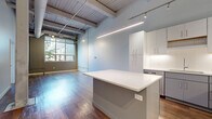 Located at 1587 E 31st St in Cleveland, OH, this 2-bedroom, 2-bathroom unit showcases a compelling blend of industrial character and modern finishes across a single floor. Soaring ceilings with exposed wooden beams, visible ductwork, and steel columns establish an unmistakable loft-style aesthetic throughout. Warm hardwood flooring flows continuously through the open-concept living and kitchen area, anchored by a large kitchen island with a crisp white quartz countertop. The kitchen is well-appointed with sleek white and gray cabinetry, a white subway tile backsplash, stainless steel appliances, and track lighting overhead. Large windows in the living area invite abundant natural light, enhancing the airy, open atmosphere. The hallway connects two generously sized bedrooms, each with access to closets—including a spacious walk-in closet with built-in shelving and clothing rods. Both bathrooms are finished with clean white vanities, large-format tile flooring, and modern fixtures. A dedicated laundry room houses a full-size washer and dryer. Transom windows above interior doors maintain the open feel while preserving privacy, making this an exceptionally well-designed urban residence.