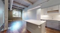 Located at 1587 E 31st St in Cleveland, OH, this 2-bedroom, 2-bathroom unit showcases a compelling blend of industrial character and modern finishes across a single floor. Soaring ceilings with exposed wooden beams, visible ductwork, and steel columns establish an unmistakable loft-style aesthetic throughout. Warm hardwood flooring flows continuously through the open-concept living and kitchen area, anchored by a large kitchen island with a crisp white quartz countertop. The kitchen is well-appointed with sleek white and gray cabinetry, a white subway tile backsplash, stainless steel appliances, and track lighting overhead. Large windows in the living area invite abundant natural light, enhancing the airy, open atmosphere. The hallway connects two generously sized bedrooms, each with access to closets—including a spacious walk-in closet with built-in shelving and clothing rods. Both bathrooms are finished with clean white vanities, large-format tile flooring, and modern fixtures. A dedicated laundry room houses a full-size washer and dryer. Transom windows above interior doors maintain the open feel while preserving privacy, making this an exceptionally well-designed urban residence.
