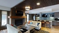 This single-floor space showcases a thoughtfully designed open-concept layout that integrates the living and kitchen areas into one cohesive environment. The living area is anchored by a contemporary linear fireplace framed in dark wood paneling, complemented by a wall-mounted flat-screen television above a warm wood mantel. A slatted wood ceiling detail adds architectural depth and a rich, modern aesthetic to this zone, contrasting elegantly with the white recessed-lit ceiling in the adjoining kitchen area.

The seating arrangement features comfortable sofas accented with colorful throw pillows, centered around a rustic wood coffee table that adds a grounding, organic element to the otherwise modern interior. Natural light filters in through a window near the fireplace wall.

The kitchen is well-appointed with dark cabinetry, a tiled backsplash in a cool blue-grey tone, stainless steel appliances including a full-sized refrigerator, and a professional-style range hood. Pendant lights hang above the kitchen island, which doubles as a breakfast bar with dark bar stools. The overall ambiance is contemporary and inviting, blending warm wood tones with modern fixtures throughout.