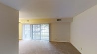 This single-floor space features an open living area characterized by neutral tones and a clean, functional layout. The room is finished with wall-to-wall carpeting in a beige tone, providing a soft, comfortable foundation throughout the space. A prominent feature of the living area is the large sliding glass door spanning much of one wall, dressed with vertical blinds that allow for flexible light control. The door provides direct access to an outdoor area, while also inviting abundant natural light into the interior. The walls are painted in a soft, warm off-white and pale yellow palette, contributing to a bright and airy ambiance. The ceiling is smooth and white, housing multiple HVAC vents that ensure comfortable climate control throughout the space. A decorative chair rail-style molding runs along one wall, adding a subtle architectural detail to the otherwise straightforward interior. The overall design is simple and understated, offering a blank canvas that is well-suited for a variety of furnishing styles. The space presents a functional, well-lit environment with practical features and a modest, uncluttered aesthetic.